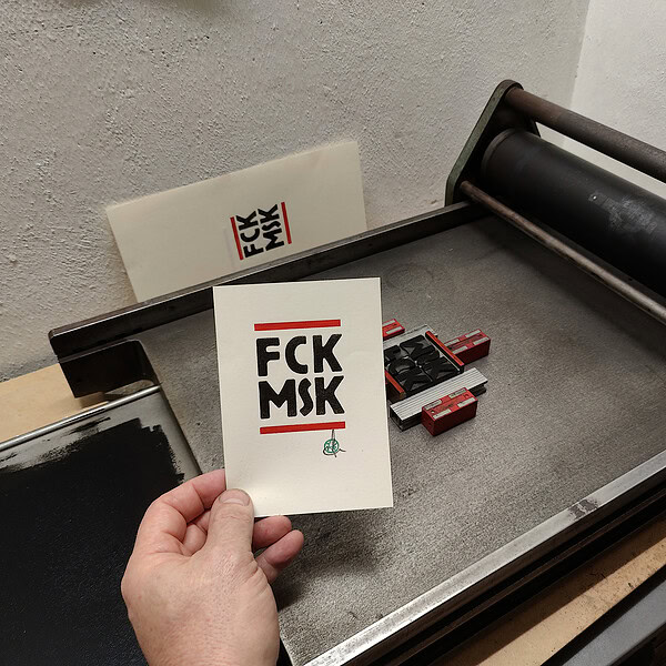 Foto meiner Hand, die eine zweifarbige Karte neben die Bleisatzform auf der kleinen Andrucknudel, einer einfachen Korrekturpresse, hält. Der schwarze Text auf der Karte lautet zwischen zwei roten Balken: FCK MSK