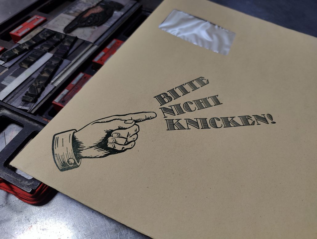 Foto eines in Bleisatz und Holz gedruckten Hinweises auf einem Großcouvert: Eine Hand zeigt auf den in drei Zeilen auffächernd gesetzten Satz: Bitte nicht knicken!