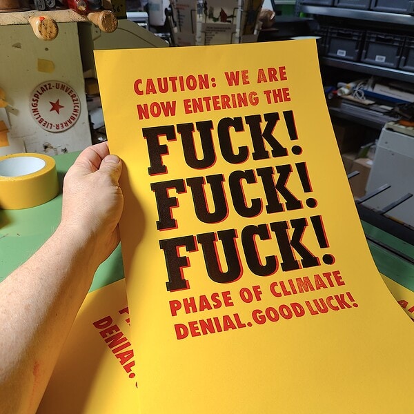Hand hält gelbes Plakat in rot und schwarz auf dem steht: Caution: We are Not entring the FUCK! FUCK! FUCK! Phase of climate denial. Good Luck!