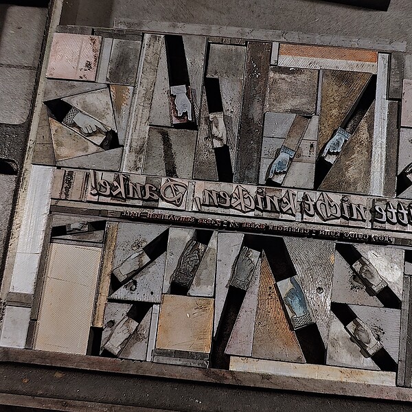 Foto einer späten Phase der Bleisatzformmontage auf dem Schließtisch. Um die Zeile Bitte nicht klicken in der Schriftart Jaguar von 1965 über der Adresszeile in der Vertigo von 1937 finden sich Setzkeile unterschiedlichster Neigungen arrangiert. Setzkeile ermöglichen das Setzen von Text oder wie hier den Schmuckhänden, Absatz der im Bleisatz üblichen Horizontalen und Vertikalen. Diese Arbeit ist puzzelig und war früher einer eigenen Gattung von Setzern vorbehalten: den Akzidenzsetzern. Hier ist die spätere Überführung der Gesamtform in ein Geviert bereits abgeschlossen, aber es fehlt noch das Fixieren der Schmuckelemente in den mit den Setzkeilen erstellten Diagonalen, das sogenannte Ausblocken.