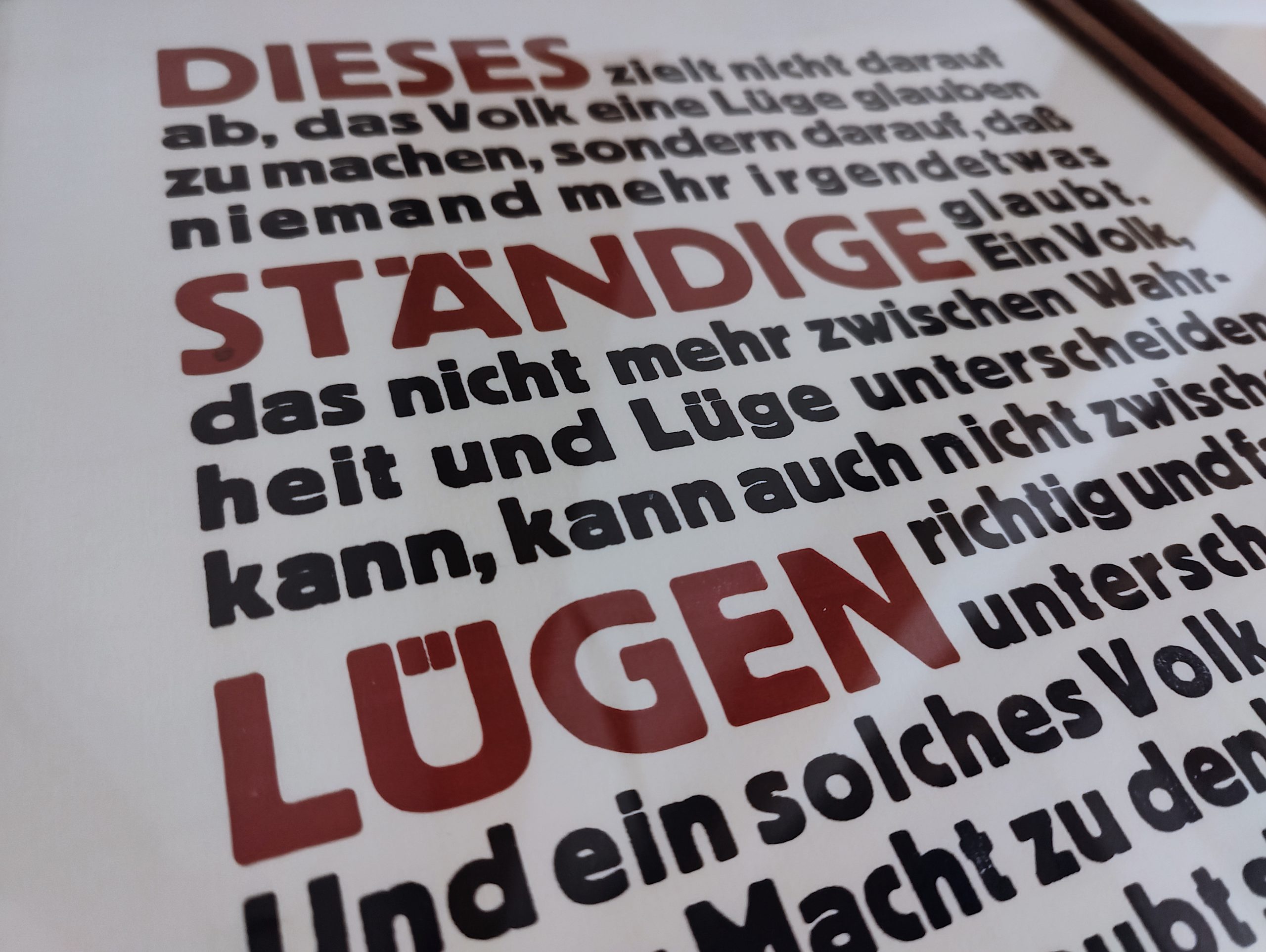 Foto Detail des fertig gerahmten Druckes in Bordaux Rot und Schwarz Blau eines Textes von Hannah Arendt. Der Text lautet: Dieses ständige Lügen zielt nicht darauf ab, das Volk eine Lüge glauben zu machen, sondern darauf, dass niemand mehr irgendetwas glaubt. Ein Volk, das nicht mehr zwischen Wahrheit und Lüge unterscheiden kann, kann auch nicht zwischen richtig und falsch unterscheiden. Und ein solches Volk, das sich seiner Macht, zu denken und zu urteilen, beraubt sieht, ist auch, ohne zu wissen und zu wollen, dem Gesetz der Lüge vollständig unterworfen. Mit einem solchen Volk kann man dann machen, was man will.