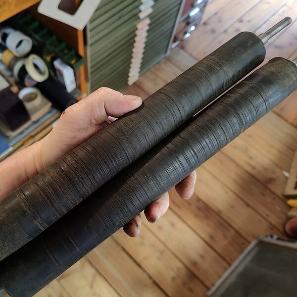 Drei schwarze, runde Holzstäbe in einem Hand gehalten, im Hintergrund Werkstattregale.