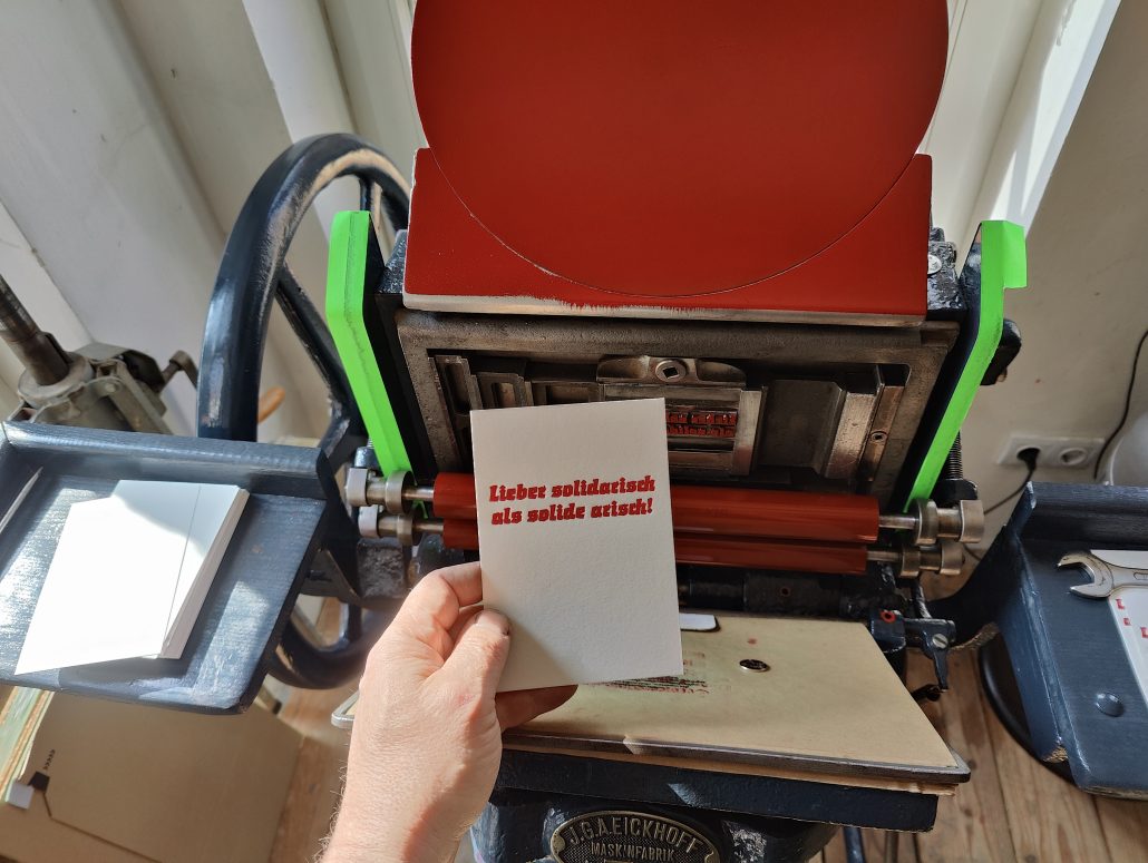 Foto meiner Hand, die eine Karte in rotem Bleisatz vor die Druckmaschine hält. Der Text der Karte: Lieber solidarisch als solide arisch! Gedruckt in der gebrochenen Werbeschrift Start von Auspurg, erschienen 1935 bei Weber.