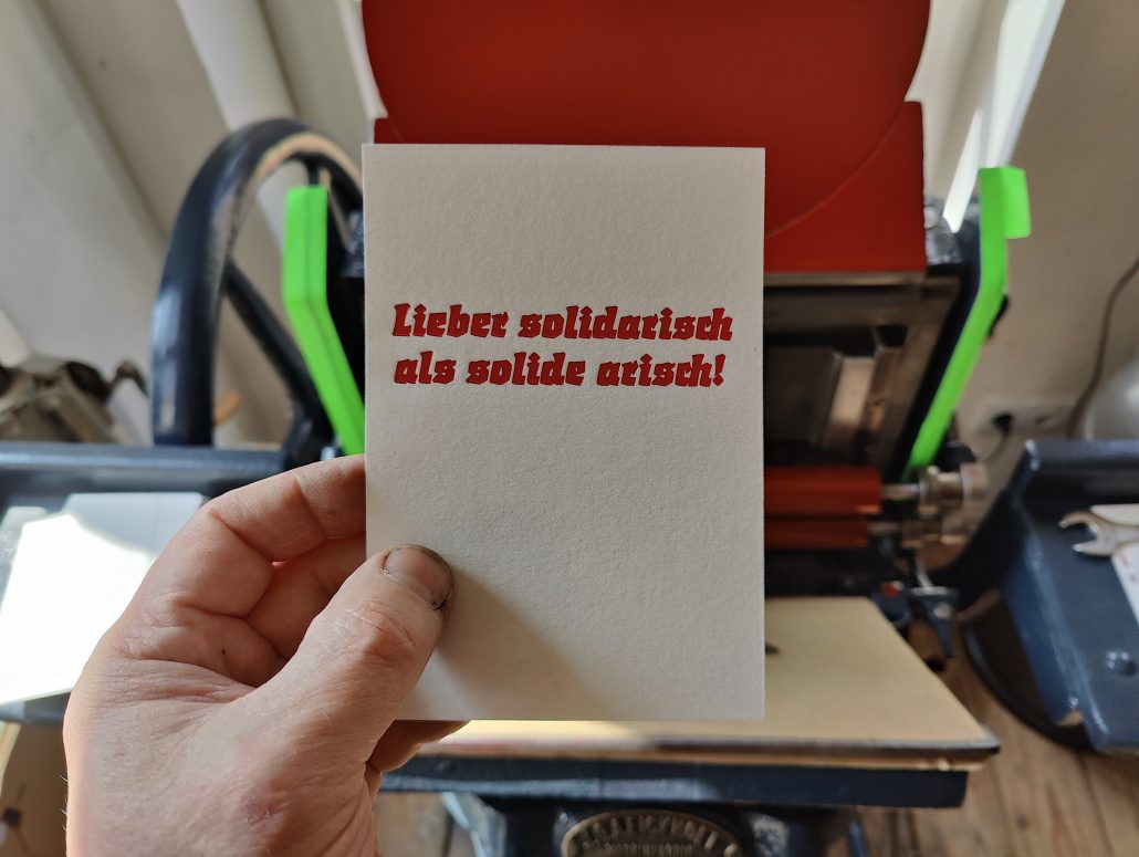 Foto meiner Hand, die eine Karte in rotem Bleisatz vor die Druckmaschine hält. Der Text der Karte: Lieber solidarisch als solide arisch! Gedruckt in der gebrochenen Werbeschrift Start von Auspurg, erschienen 1935 bei Weber.