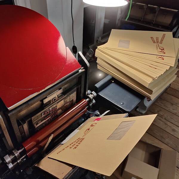 Foto einer Situation beim Bedrucken von C4-Versandtaschen am Tret-Tiegel, einer 120 jähre alten Druckmaschine, die mit Fußbetrieb läuft.