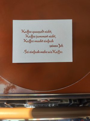 Foto meiner Hand, die eine in braun gedruckte Karte in Bleisatz vor die Druckmaschine hält. Der Text der in einer typischen 1950er Eisdielen und Caféterassen-Werbeschreibschrift ausgeführt ist lautet: Kaffee quasselt nicht, Kaffee jammert nicht, Kaffee macht einfach seinen Job. -Sei einfach mehr wie Kaffee.