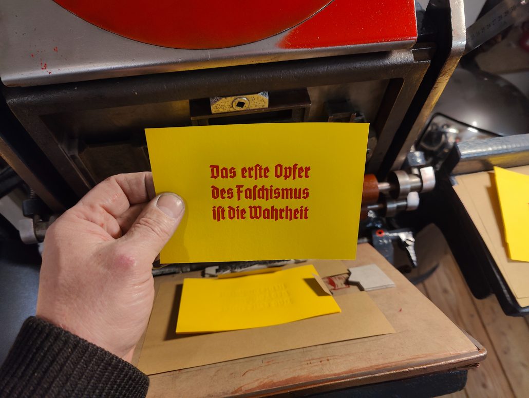 Foto meiner Hand die einen roten Abzug auf gelben Grund vor eine Tiegeldruckpresse hält. Der Text in einer Neotextura, also einer Mischung aus umgangssprachlich Frakturschrift und moderner Grotesk lautet: Das erste Opfer des Faschismus ist die Wahrheit.