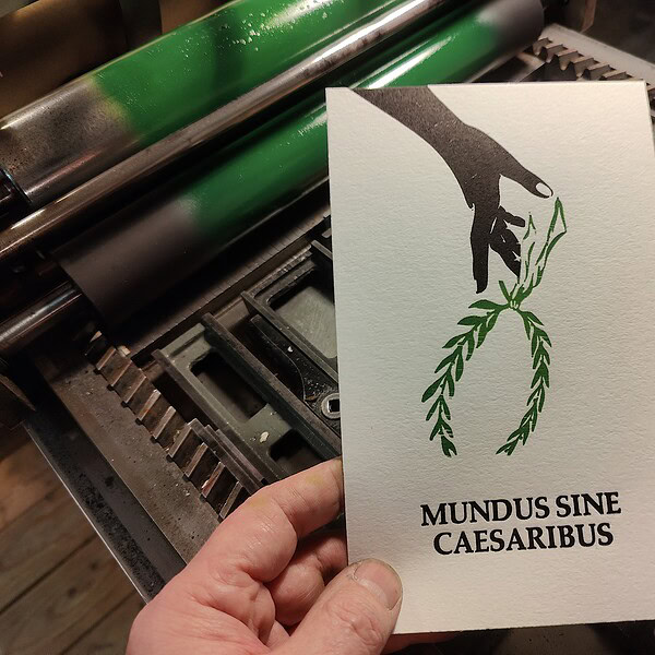 Foto: Hand hält Karte vor Druckmaschine. Zu sehen ist ein zweifarbiger Abzug - eine einen über den Handrücken nach unten geöffnete Hand, die einen Lorbeerkranz fallen lässt über dem Schriftzug Mundus Sine Caesarius. Der Abzug ist auf Baumwollpapier.