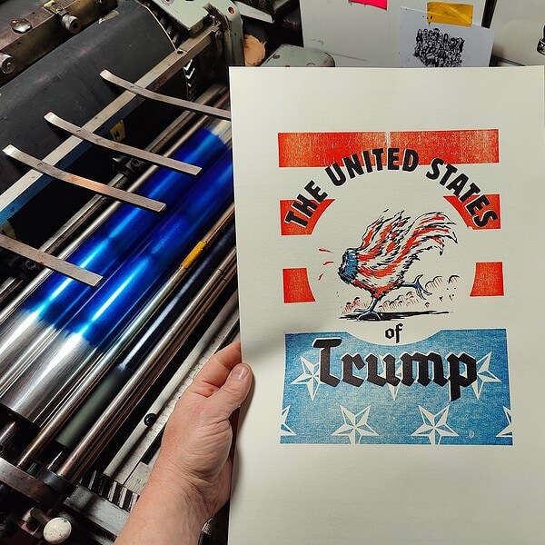 Foto meiner Hand, die ein dreifarbiges Plakat eines kopflos rennenden Gockels in Farben der Vereinigten Staaten von Amerika umrundet vom Schriftzug The United Staates of Trump vor die Druckmaschine hält