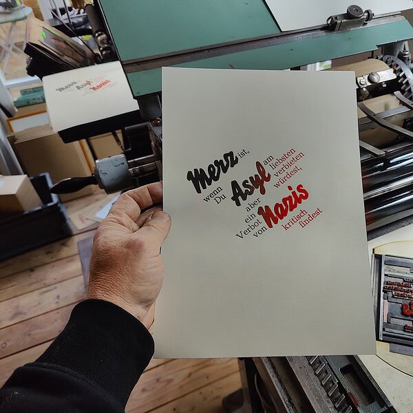 Foto von Rot-schwarzem Irisdruck neben der Form auf der Druckmaschine. Der Text lautet: Merz ist, wenn Du Asyl am liebsten verbieten würdest, aber ein Verbot von Nazis kritisch findest