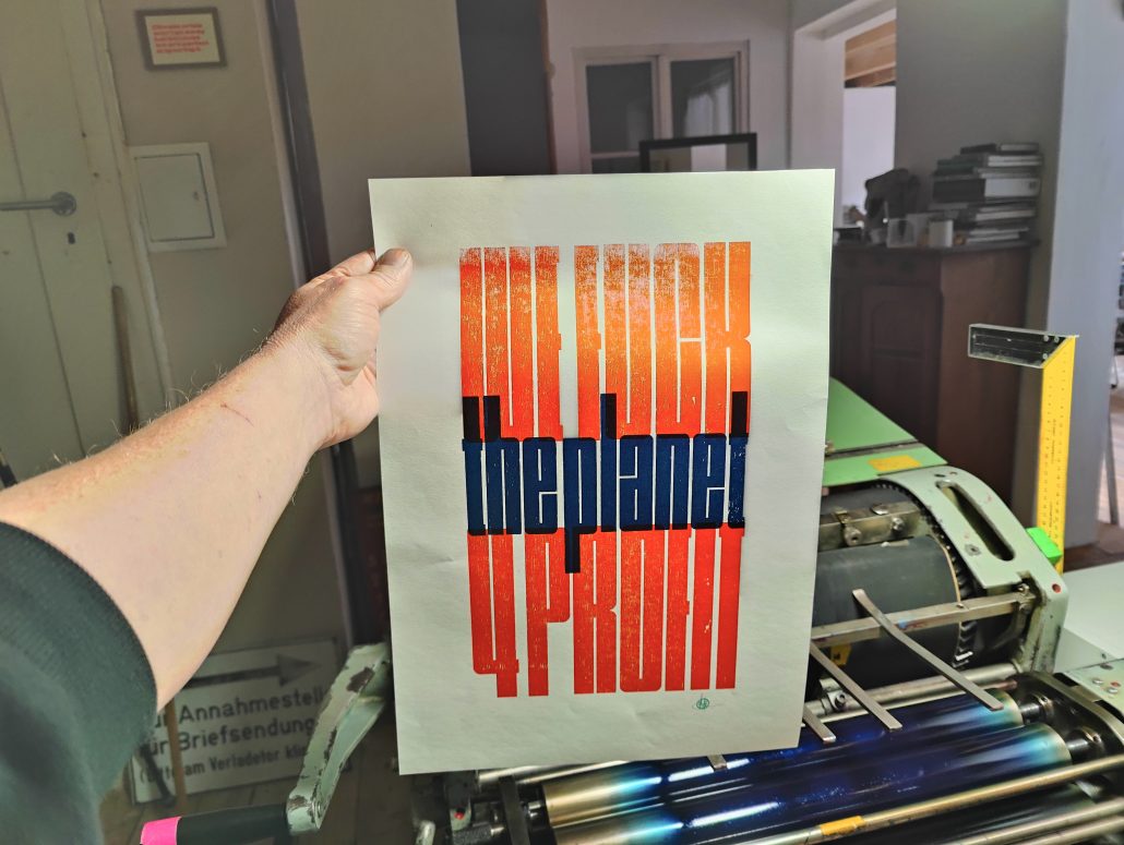 Foto von einem zweifarbigen Druck neben der zweiten Form auf der Druckmaschine. der graphische, sehr enge, schmalhohe Text lautet:
WE FUCK
the planet
4 PROFIT
die Großbuchstanben sind rotorange, die Kleinbuchstaben dunkelblau. 