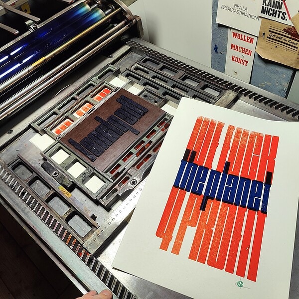 Foto von einem zweifarbigen Druck neben der zweiten Form auf der Druckmaschine. der graphische, sehr enge, schmalhohe Text lautet: WE FUCK the planet 4 PROFIT die Großbuchstanben sind rotorange, die Kleinbuchstaben dunkelblau.