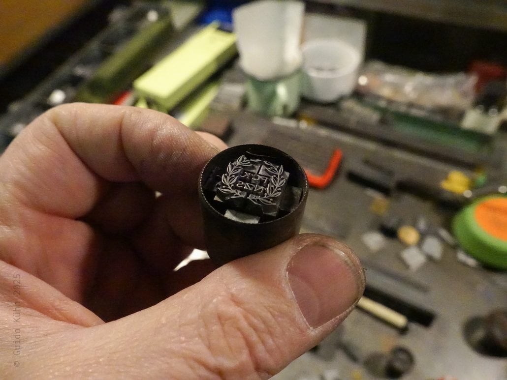 Hand hält eine schwarze Zündkerze mit Logo, Werkstatt im Hintergrund.