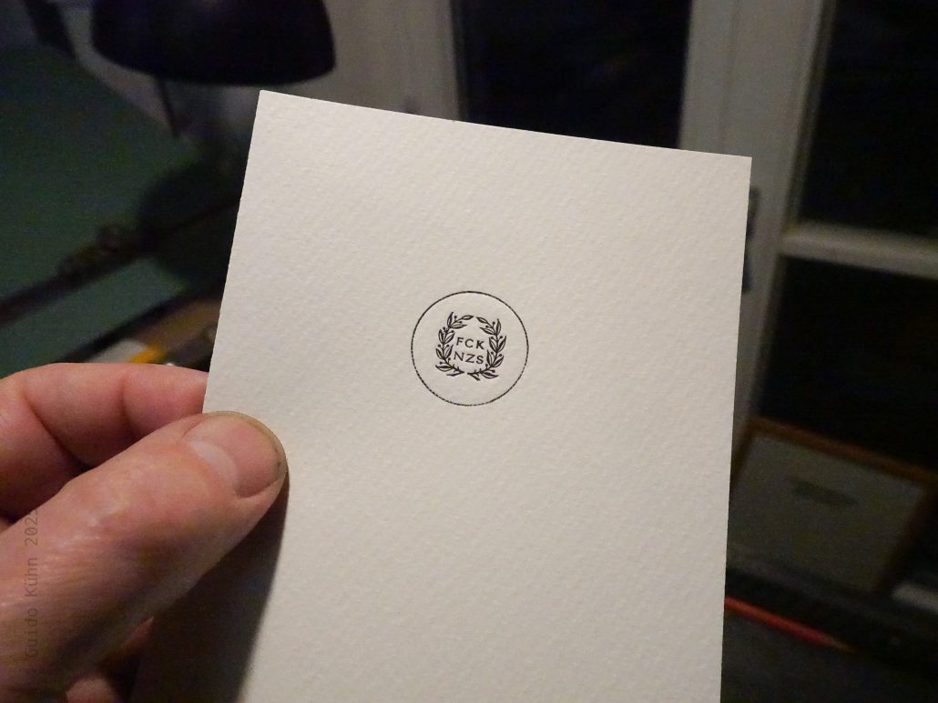 Hand hält eine weiße Karte mit FCK NZS Logo in Innenraum.