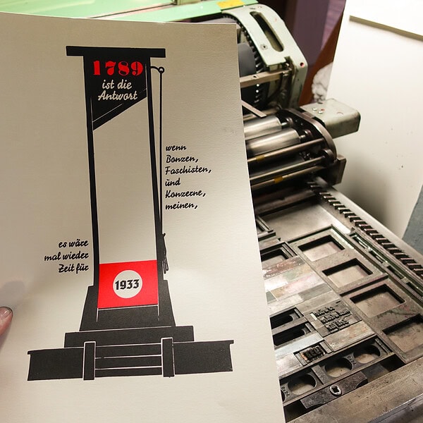 Foto des fertigen Druckes. Zu sehen ist eine schwarze Silhouette einer Guillotine mit der rot hinterdruckten Jahreszahl 1728 in der alten Antiqua Normande, darunter invers in der Signal der Teilsatz "ist die Antwort" seitlich rechts steht die Fortsetzung "wenn Bonzen, Faschisten und Konzerne meinen," darunter seitlich links der Guillotine "es wäre mal wieder Zeit für" und im im roten Kopfschild die Jahreszahl 1933 in der Tannenberg Fraktur als Abschluss des Satzes. Das Papier ist aus drucktechnischen Gründen noch unbeschnitten, daher ist das Format noch länger