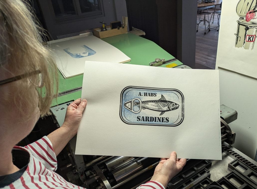 Foto einer Frau, die einen fertigen Druck in die Kamera hält. Das Motiv ist ein zweifarbiger Holzschnitt einer Sardinendose, auf der eine Sardine über und unter der der Schriftzug A. HABS SARDINEN zu lesen ist. 