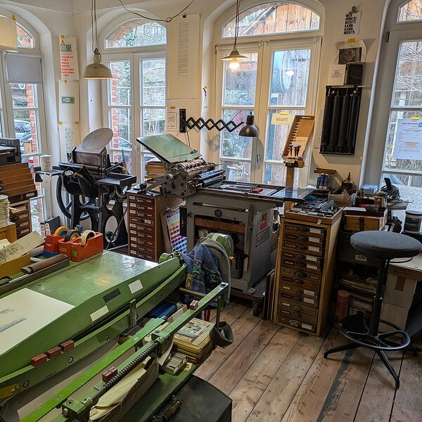 Ein heller, handwerklich geprägter Druckereiraum voller traditioneller Drucktechnik. Mehrere historische Druckpressen stehen dicht beieinander, darunter eine kleine Zylinderpresse und eine zwei Tiegelpressen. Links und rechts reihen sich hohe Setzkästen und Schubladenschränke, gefüllt mit Bleisatz, Holzlettern und Werkzeugen. Auf den Arbeitsflächen liegen Papierstapel, Druckbögen und verschiedene Hilfsmittel. Im Vordergrund steht eine grüne Schneidemaschine, auf deren Fläche frisch gedruckte Blätter liegen. Die Wände sind mit typografischen Postern und Informationsblättern dekoriert. Durch große Rundbogenfenster fällt warmes Tageslicht in den Raum und beleuchtet die etwas chaotisch anmutende, aber sorgfältig organisierte Werkstatt