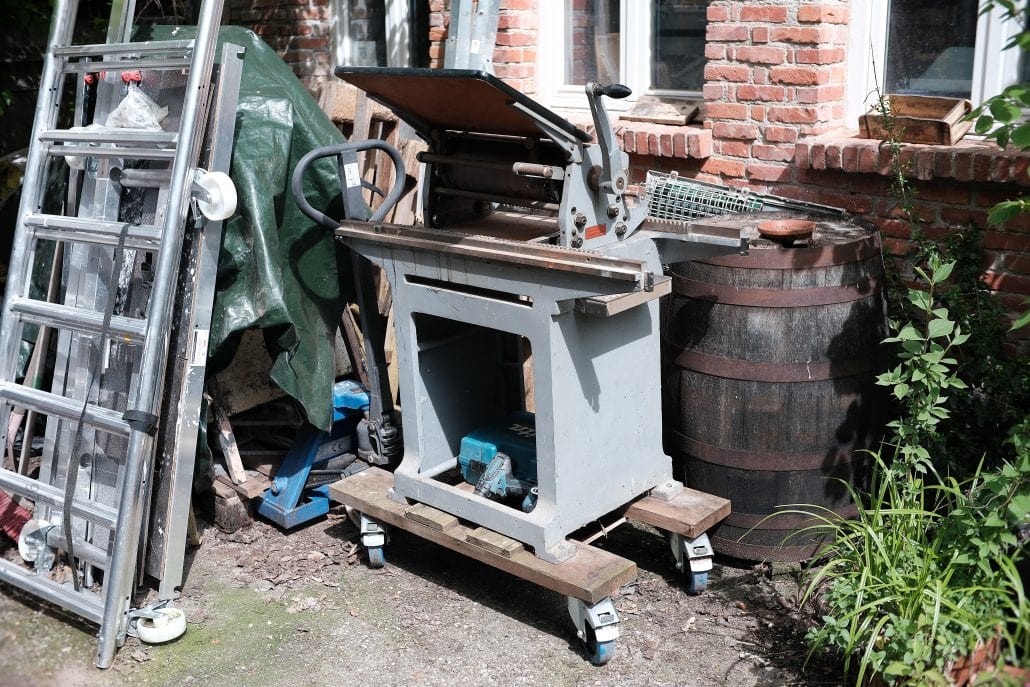 Alte Tischkreissäge im Garten neben einem Weinfass und Pflanzen.
