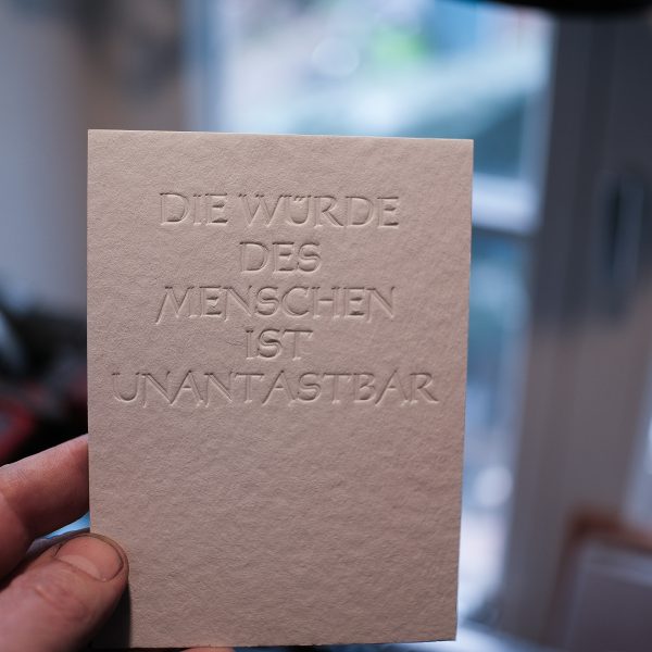 Foto meiner Hand, die einen Büttenkartonn in 600g mit einer Blindprägung in der 1950er Urkunden Schriftart Erler Versalien hochhält. Der Text lautet: DIE WÜRDE DES MENSCHEN IST UNANTASTBAR.