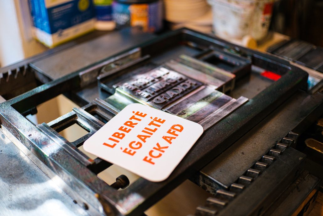 ViereckigerBierdeckel mit dem in kapitaler Futura gesetztem und signalleuchtorange gedrucktem auf Block gesetztem Dreizeiler: LIBERTÉ ÉGALITÉ FCK AFD neben der Bleisatzform